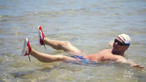 Yüzme şortu, güneş gözlüğü ve blazer bir adam sığ suda yatıyor, su sıçrayan, güneş ışınları. Sıcak bir yaz gününde güneşlenme, denizde tatil. — Stok video