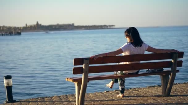 Été, coucher de soleil, remblai au bord de la mer, une jeune femme brune assise sur un banc, dos, admirant un beau coucher de soleil. femme seule sur un banc au bord de la mer au coucher du soleil — Video