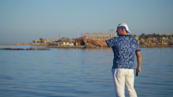 Un homme sur le rivage montre quelque chose à la main aux gens sur un yacht en mer. homme sur la plage agitant les bras. baie maritime, marina, en été au coucher du soleil . — Video