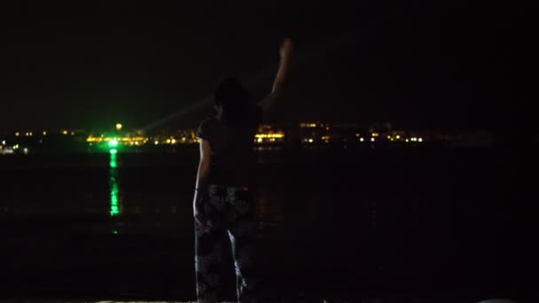 Noc, Kobieta tańczy, widok z tyłu. przed światłami świecącego miasta nad wodą — Wideo stockowe