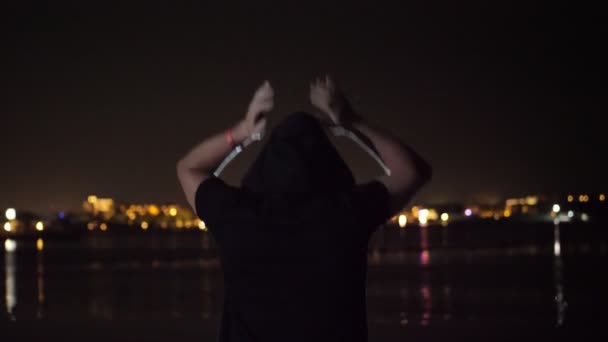 Noche, un hombre en una capucha negra está bailando, vista desde atrás. contra las luces de la ciudad resplandeciente sobre el agua — Vídeos de Stock