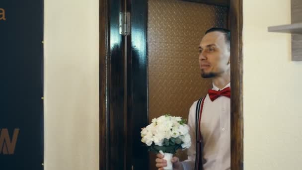 En la puerta entra el joven, el novio, con una pajarita roja y con un ramo de flores. mira a la novia . — Vídeo de stock