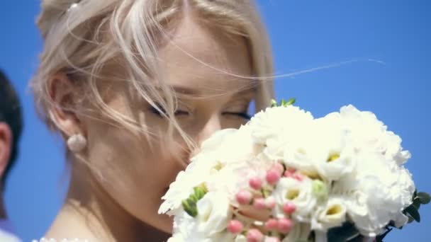 Portret, close-up, mooie blonde bruid genieten van de geur van haar bruiloft boeket van witte bloemen. haar blonde krullen fladderen in de wind. tegen de blauwe lucht. zonnige dag in de zomer. Bruiloft — Stockvideo