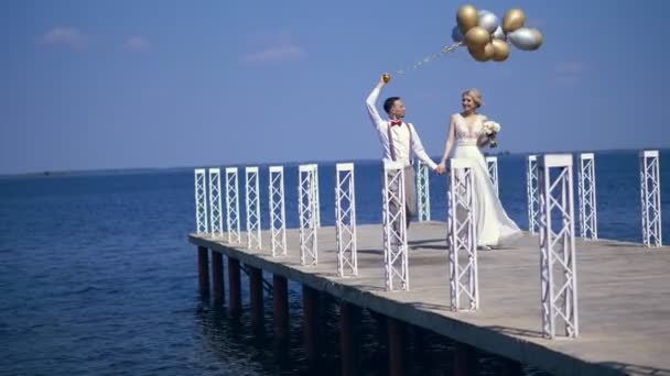 Giovani, bella coppia sposi stanno camminando sul bellissimo molo, lo sposo tiene in mano palloncini, guardarsi, sorridendo, contro il mare blu e il cielo. giornata estiva di sole. matrimonio — Video Stock