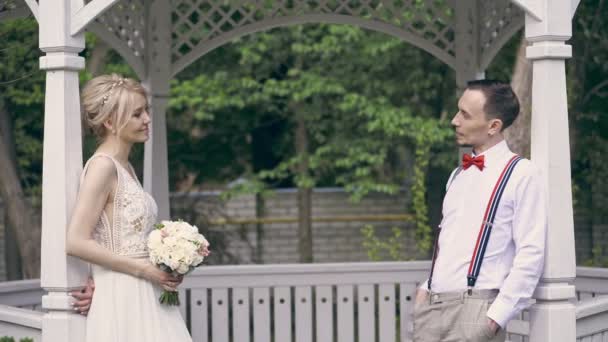 Ein junges schönes Paar steht sich in der Laube gegenüber und reicht einander einen Brautstrauß, albert herum und lächelt. Sonniger Frühlingstag. Hochzeit — Stockvideo