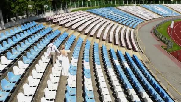 空中视频，在体育场，身着婚纱的新婚夫妇在白色和蓝色的座位之间行走。春天阳光明媚的日子。婚礼 — 图库视频影像