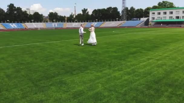 Video aereo, allo stadio, al centro di un campo di calcio verde, sposi felici in abito da sposa stanno correndo attraverso il campo di calcio. giornata di sole primaverile. matrimonio — Video Stock