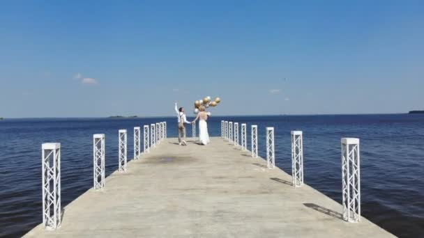 Giovani, bella coppia sposi stanno camminando sul bellissimo molo, lo sposo tiene in mano palloncini, guardarsi, sorridendo, contro il mare blu e il cielo. giornata estiva di sole. matrimonio — Video Stock