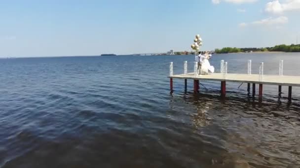 航空ビデオ、美しい新婚夫婦、新郎新婦が屋外で踊る、美しい桟橋で、青い海と空に対して。新郎は、彼の手に風船を保持します, 夏の晴れた日.結婚 式 — ストック動画