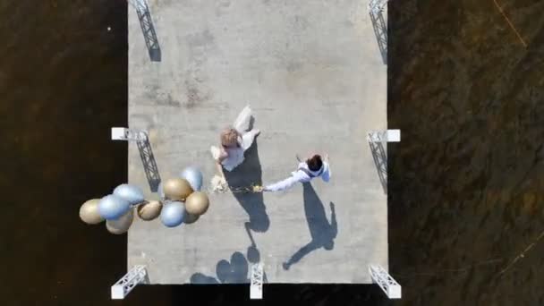 Vídeo aéreo, vista superior, hermosa pareja recién casada, novia y novio bailando al aire libre, en un muelle, novio sostiene globos en su mano, día soleado de verano. boda — Vídeos de Stock