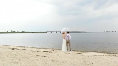 aero, güzel yeni evliler sahilde duran, şeffaf bir şemsiye altında, mavi gökyüzü, nehir ve nehir üzerinde büyük bir köprü karşı. bahar güneşli ve biraz yağmurlu bir gün. Düğün.