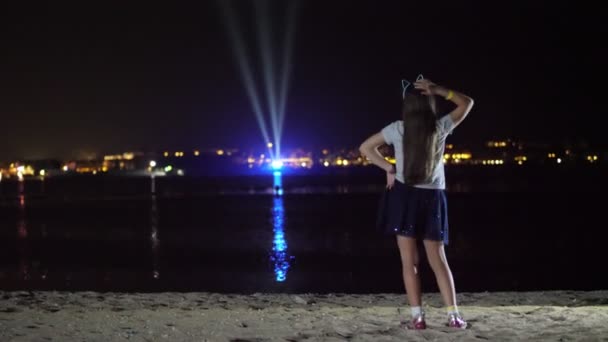 Noc, Teen Girl w błyszczącej spódnicy tańczy, widok z tyłu. przed światłami świecącego miasta nad wodą — Wideo stockowe