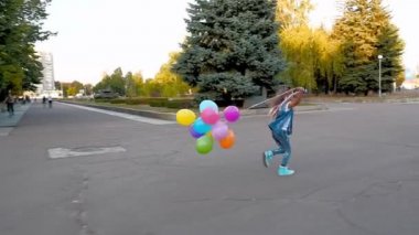 Kot pantolonlu genç bir kız elinde bir sürü renkli balonla parkta koşuyor..