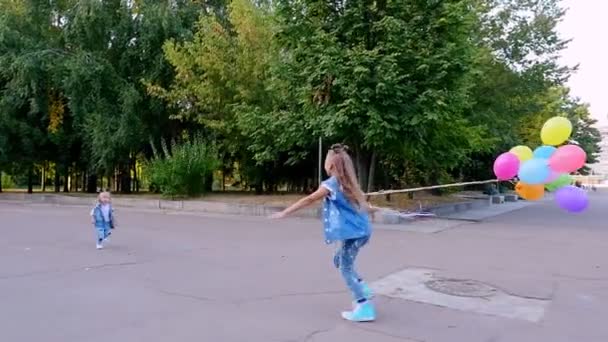 Duas meninas irmãs, uma adolescente e uma pequena correm uma para a outra no parque, com um grande bando de balões coloridos. Eles estão abraçando — Vídeo de Stock