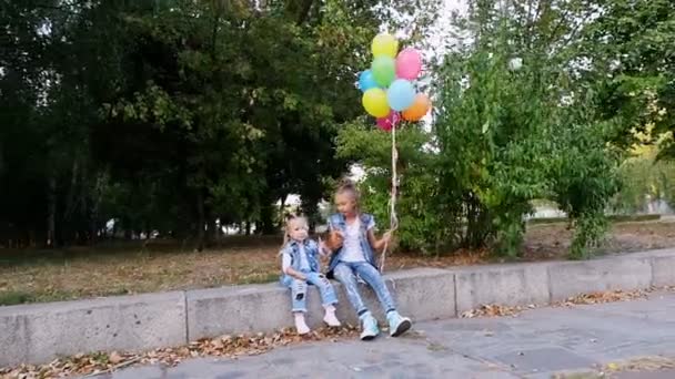 Twee kleine meisjes zusjes, een tienermeisje en een kleintje zittend op een bankje in een park, met een grote bos kleurrijke ballonnen. Ze praten. — Stockvideo