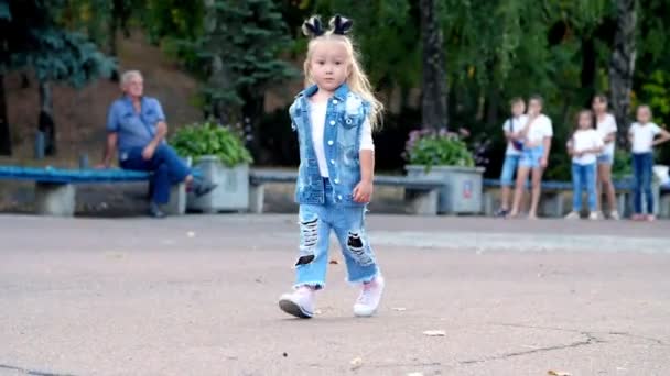 En liten flicka i en stilren jeansdräkt och hästsvans på huvudet går genom parken. — Stockvideo
