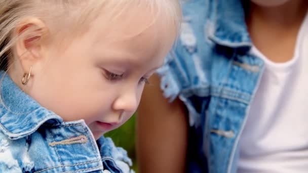 Portret, twee kleine meisjes zusjes in jeans pakken, een tienermeisje en een kleintje zitten op een bankje in een park, met een grote bos kleurrijke ballonnen. Ze praten. — Stockvideo