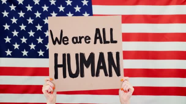 Manifestante sostiene una pancarta con un lema - TODOS somos HUMANOS - en el contexto de la bandera de Estados Unidos. Lucha contra el racismo, por la igualdad de derechos en Estados Unidos . — Vídeos de Stock