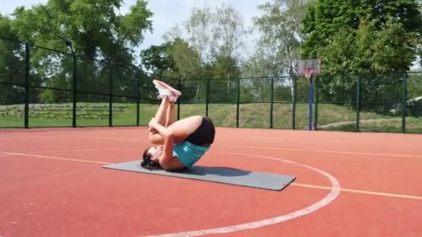 Фітнес-леді в спортивному одязі роблять різні вправи на спортивному килимку, на помаранчевому баскетбольному полі з білими позначками, на стадіоні. Спорт на відкритому повітрі. онлайн-тренінги. літній день . — стокове відео