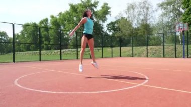Spor kıyafetli kadın, stadyumdaki beyaz işaretli turuncu bir basketbol sahasında çeşitli egzersizler yapıyor. Açık hava sporları. Online eğitim. yaz günü.
