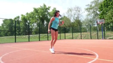 Spor kıyafetli kadın, stadyumdaki beyaz işaretli turuncu bir basketbol sahasında çeşitli egzersizler yapıyor. Açık hava sporları. Online eğitim. yaz günü.