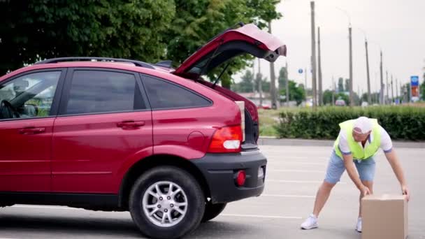 Corriere maschio, in un gilet speciale, berretto e pantaloncini, scarico pacchi, scatole di cartone dal bagagliaio dell'auto, all'aperto. Servizio di consegna merci. — Video Stock