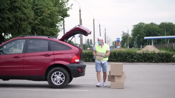 Mensageiro masculino, em um colete especial, máscara protetora, luvas, pacotes de carregamento, colocando caixas de papelão no porta-malas do carro, serviço de entrega de carga em quarentena, surto de coronavírus . — Vídeo de Stock