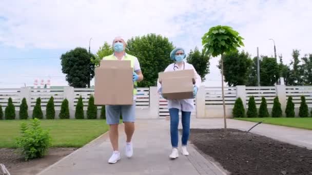 Courier and doctor, in protective masks, gloves, going through courtyard of hospital or medical facility, carrying boxes with medical equipment, during coronavirus outbreak, quarantine. — Stock Video
