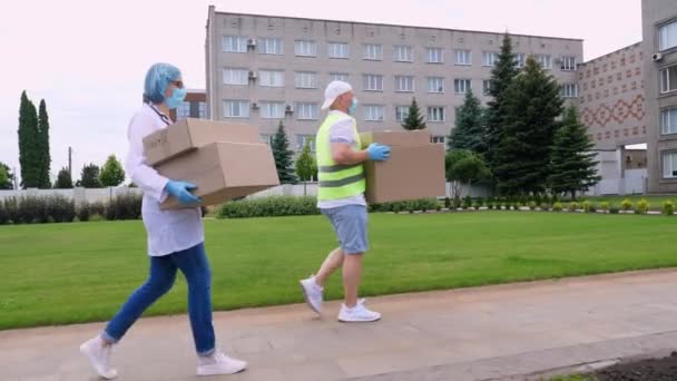 Courier and doctor, in protective masks, gloves, going through courtyard of hospital or medical facility, carrying boxes with medical equipment, during coronavirus outbreak, quarantine. — Stock Video