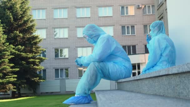 Exhausted and tired doctor and nurse in protective uniforms, masks, glasses, are sitting on floor in courtyard of hospital or medical facility. Coronavirus outbreak. — Stock Video