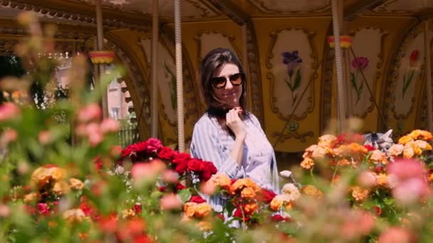 Schöne brünette Frau, in Sonnenbrille, posiert neben einem Blumenbeet mit Rosen, in einem Park, auf dem Hintergrund des Karussells. Sommersonniger Tag im Freizeitpark. — Stockvideo
