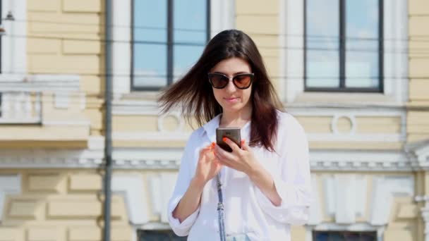 Güneş gözlüklü güzel esmer kadın cep telefonunu ve akıllı telefonunu kullanıyor. Açık havada, eski şehir binasının arka planına karşı. Yaz güneşli bir gün — Stok video