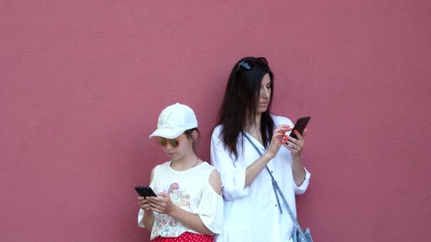 Bella donna bruna e una bambina stanno usando i loro telefoni cellulari, smartphone. all'aperto, sullo sfondo della parete rosa scuro. estate giornata di sole — Video Stock