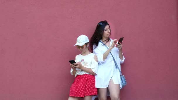 Mooie brunette vrouw en een kind-meisje gebruiken hun mobiele telefoons, smartphones. buiten, tegen de achtergrond van donkerroze wand. zonnige zomerdag — Stockvideo