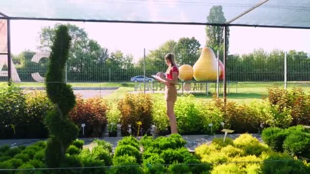 Γυναίκα κηπουρός ελέγχει την ποιότητα των φυτών, κρατά σημειώσεις με στυλό στο σημειωματάριο, στο κέντρο του κήπου ή το θερμοκήπιο. καλλιέργεια και φροντίδα των φυτών. ανθοκομία, κηπουρική, κηπουρική, ανθοκομία. — Αρχείο Βίντεο