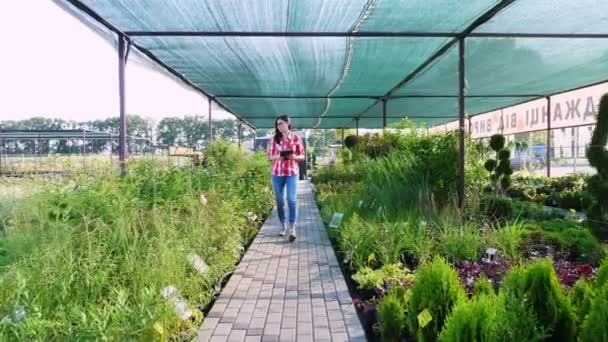 Il giardiniere femminile controlla la qualità di impianti, prende appunti su targa digitale, camminando lungo centro di giardino o serra. crescere e prendersi cura delle piante. floristica, giardinaggio, orticoltura, floricoltura. — Video Stock