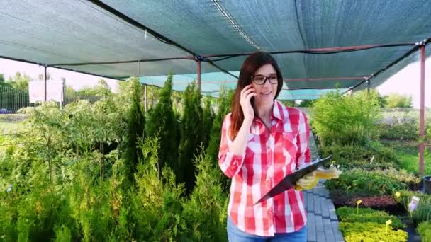 Γυναίκα κηπουρός ή ανθοπώλης μιλάει στο τηλέφωνο, κρατώντας ψηφιακή ταμπλέτα, στο κέντρο κήπου ή στο θερμοκήπιο. καλλιέργεια και φροντίδα των φυτών. ανθοκομία, κηπουρική, κηπουρική, ανθοκομία. — Αρχείο Βίντεο
