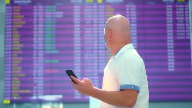 Maskeli erkek yolcu, havaalanında kalkış panosuna karşı duruyor. Uçuş saatlerini cep telefonuyla kontrol ediyor. Coronavirüs salgınından sonra hava yolculuğu açılıyor..