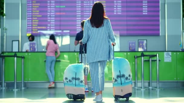 航空旅客、女性の観光客は、荷物を持ち、空港で出発ボードに対して立っている。彼女は飛行機の時刻表を調べている。コロナウイルス流行後の航空旅行の開始 — ストック動画