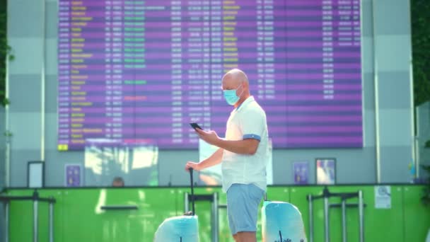 Mannelijke vliegtuigpassagier met medisch masker, met bagage, staande tegen het vertrekbord op de luchthaven. hij controleert de dienstregeling van de vlucht met mobiel. opening van het luchtverkeer na de coronavirusepidemie. — Stockvideo