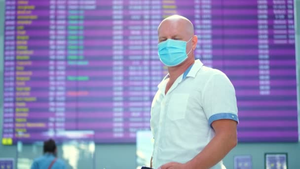 Portret van een man met een medisch masker op de luchthaven. Vliegtuigpassagier, toerist staat tegen vertrekdienstregeling. opening van het luchtverkeer na de coronavirusepidemie. — Stockvideo