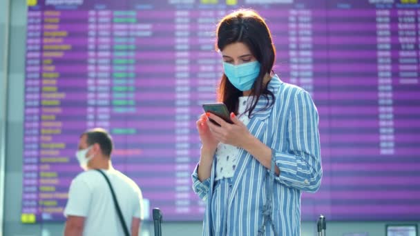 Passagère en masque, debout contre le panneau de départ à l'aéroport. elle vérifie l'horaire des vols à l'aide du mobile. ouverture du voyage aérien après l'épidémie de coronavirus. — Video