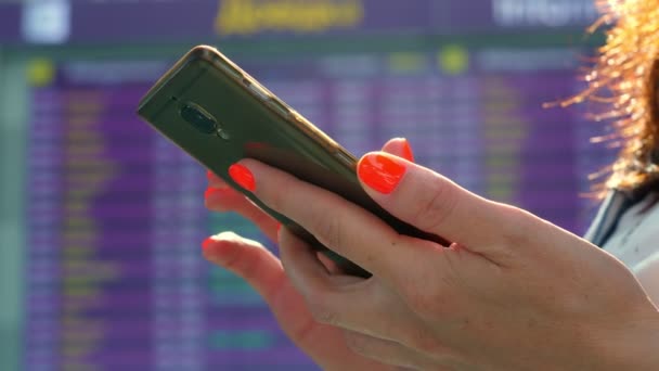 Primer plano, manos femeninas utilizando móvil, en el fondo de la tabla de horarios de vuelo de salida, en el aeropuerto. — Vídeo de stock