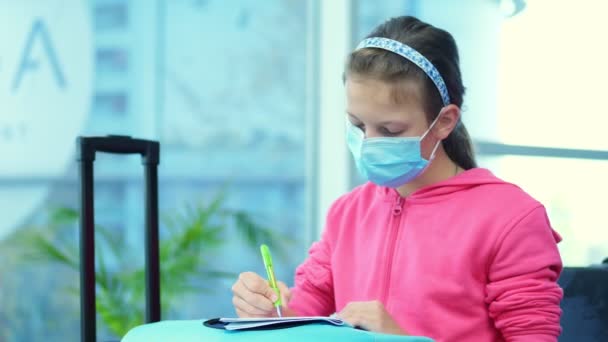 Retrato de una chica, niño con máscara médica, dibujando un cuaderno en una maleta, equipaje, en el aeropuerto. esperando la salida. apertura de los viajes aéreos tras la epidemia de coronavirus. — Vídeos de Stock