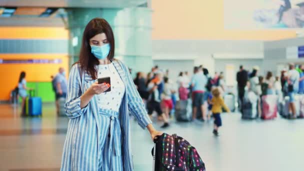 Pasajera femenina en máscara, de pie en el aeropuerto, con equipaje, esperando para abordar el avión. está comprobando el horario de vuelo usando el móvil. apertura de los viajes aéreos tras la epidemia de coronavirus. — Vídeo de stock