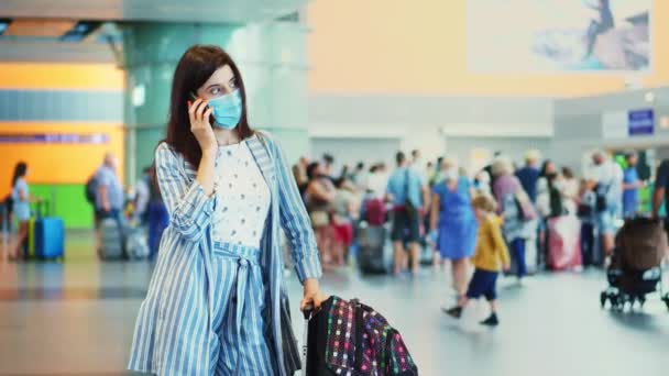 Жінка в масці, розмовляє на мобільному телефоні, на тлі натовпу пасажирів повітря, в аеропорту. очікування на літак з багажем. відкриття повітряних подорожей після епідемії коронавірусу . — стокове відео