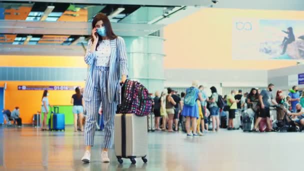 空港で乗客の群衆を背景に携帯電話で話している女性です。荷物を持って飛行機に乗るのを待ってる。コロナウイルス流行後の航空便の開設. — ストック動画