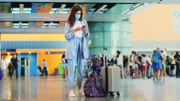 Maskeli kadın, havaalanında duruyor, bavuluyla uçağa binmeyi bekliyor. Uçuş saatlerini cep telefonuyla kontrol ediyor. Coronavirüs salgınından sonra hava yolculuğu açılıyor.. — Stok video