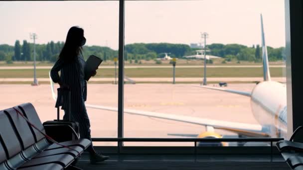 Tmavá silueta obchodnice v ochranné masce, s kufříkem, příručním zavazadlem a laptopem v rukou, kráčející proti velkému oknu v čekárně na letišti. otevření letecké dopravy — Stock video