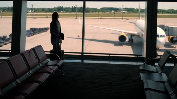 Dunkle Silhouette einer Geschäftsfrau mit Koffer und Laptop in der Hand, die gegen ein großes Fenster im Wartezimmer der Duty Free Zone am Flughafen läuft. — Stockvideo